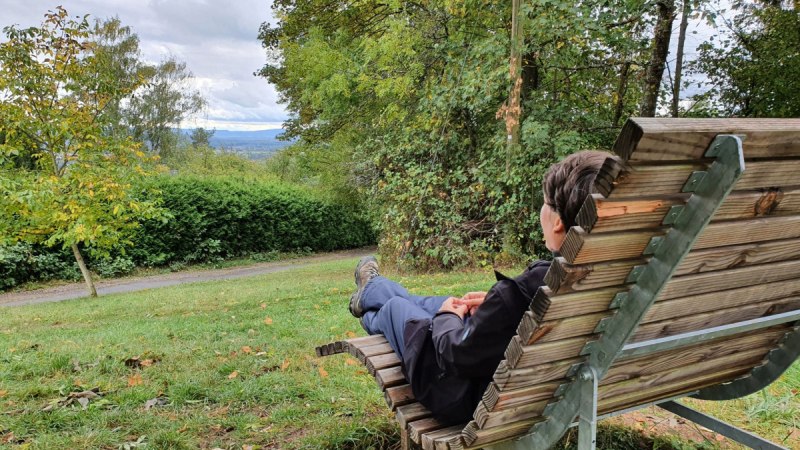 Eine Person sitzt entspannt auf einer Holzbank und blickt auf eine gr&uuml;ne, bewaldete Landschaft mit bew&ouml;lktem Himmel.