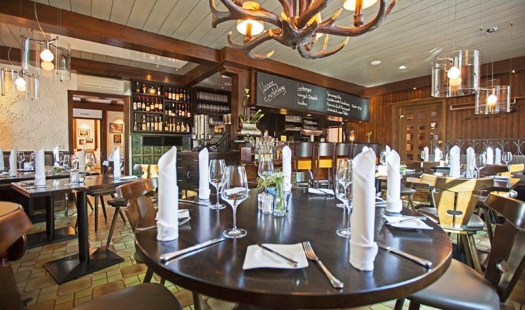 Ein stilvolles Restaurant mit runden Tischen, elegant gedeckt mit Weingläsern und Servietten. Ein Kronleuchter aus Geweihen hängt von der Decke., © Hotel Waldhorn Ein stilvolles Restaurant mit runden Tischen, elegant gedeckt mit Weingläsern und Servietten. Ein Kronleuchter aus Geweihen hängt von der Decke., © Hotel Waldhorn