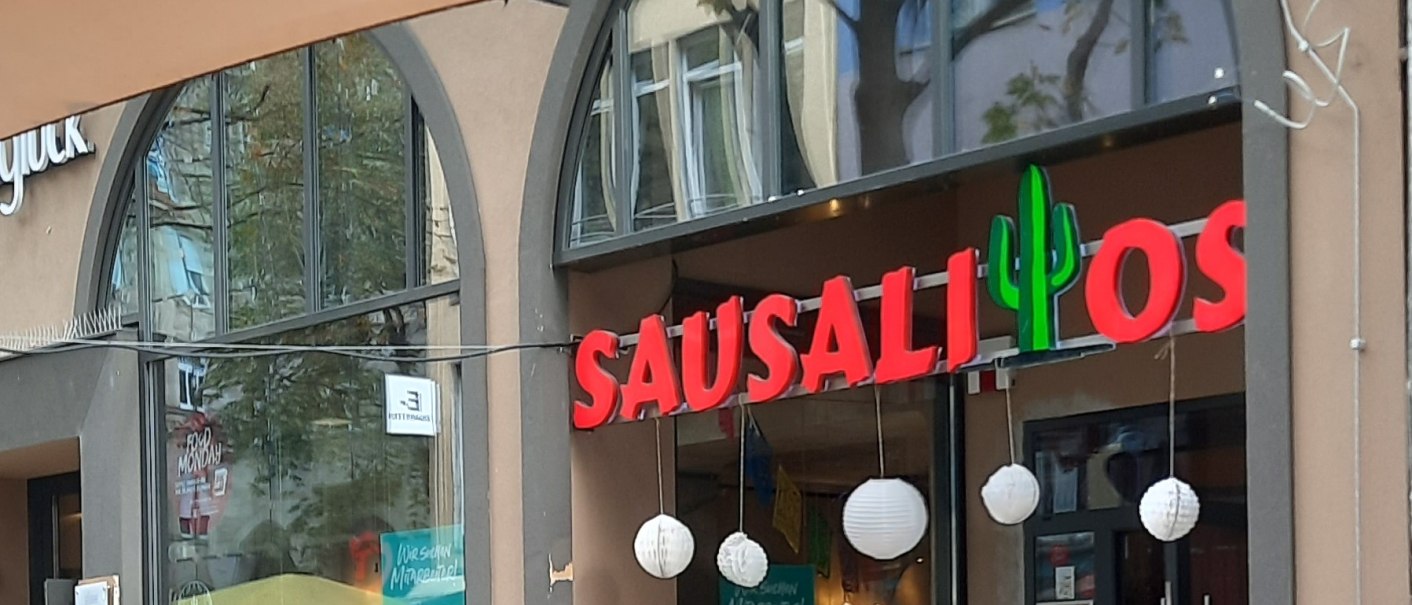 Eingang des Restaurants Sausalitos mit rotem Schriftzug und Kaktus-Dekoration. Außenbereich mit Holztischen und Werbetafel für Taco Tuesday., © SMG Eingang des Restaurants Sausalitos mit rotem Schriftzug und Kaktus-Dekoration. Außenbereich mit Holztischen und Werbetafel für Taco Tuesday., © SMG