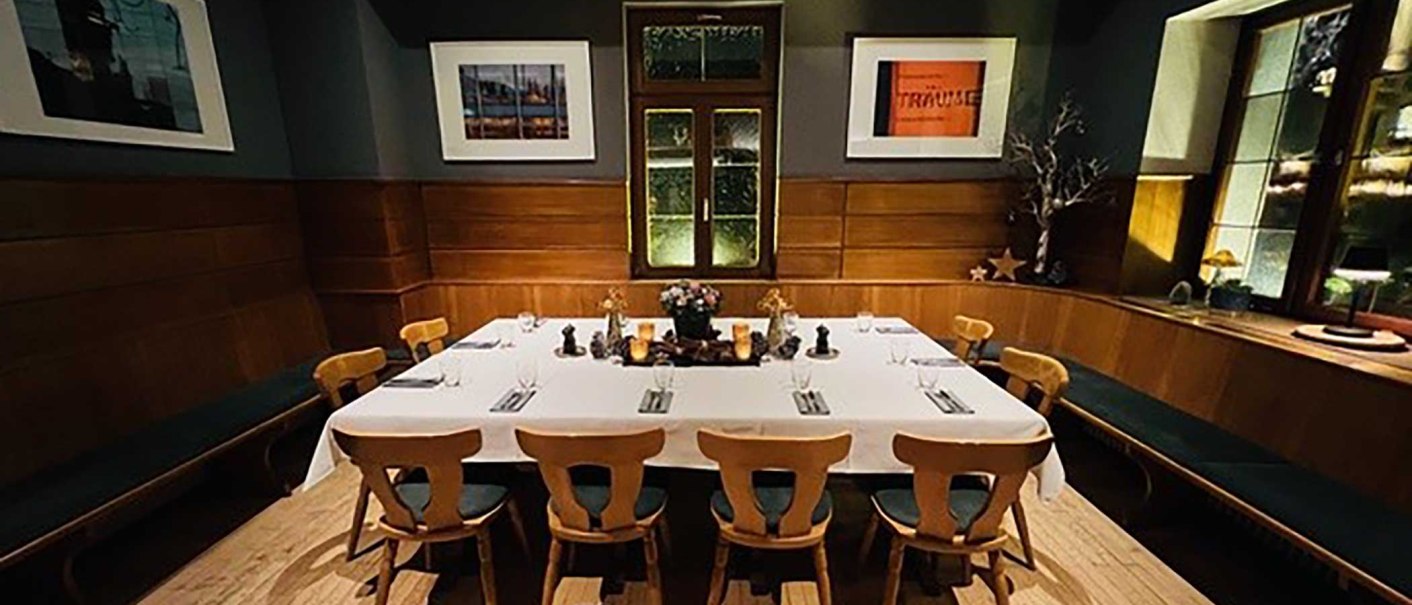 Ein eleganter Raum mit Holzwänden, einem großen, weiß gedeckten Tisch und Stühlen. An den Wänden hängen Kunstwerke, und das Fenster ist dekorativ gestaltet., © Zumbecher Ein eleganter Raum mit Holzwänden, einem großen, weiß gedeckten Tisch und Stühlen. An den Wänden hängen Kunstwerke, und das Fenster ist dekorativ gestaltet., © Zumbecher