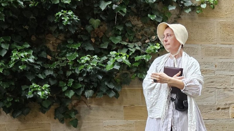 Eine Frau in historischer Kleidung steht nachdenklich vor einer mit Efeu bewachsenen Steinmauer. Sie h&auml;lt ein Buch in der Hand., &copy; Xenia Busam