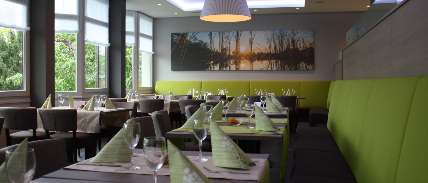 Modernes Restaurant mit gedeckten Tischen, grünen Servietten und großen Fenstern. An der Wand hängt ein Bild mit Sonnenuntergangsmotiv., © Hotel am Schlossberg Herrenberg Modernes Restaurant mit gedeckten Tischen, grünen Servietten und großen Fenstern. An der Wand hängt ein Bild mit Sonnenuntergangsmotiv., © Hotel am Schlossberg Herrenberg