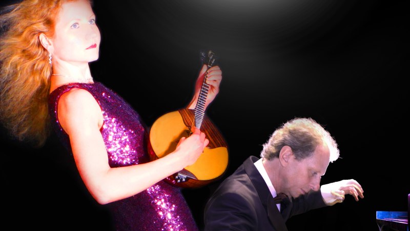 Eine Frau in einem glitzernden Kleid spielt eine Domra, während ein Mann im Anzug Klavier spielt. Der Hintergrund ist schwarz mit einem Lichtschein., © DomraPiano