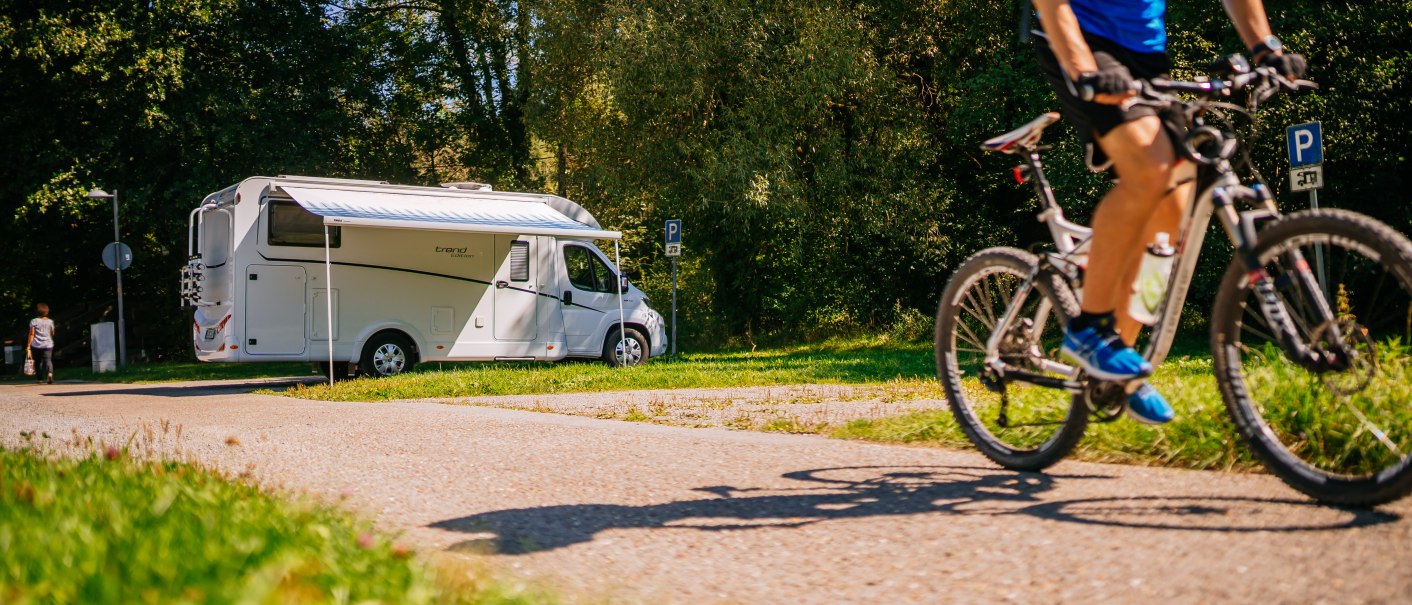 Wohnmobil auf Stellplatz in gr&uuml;ner Umgebung, Radfahrer f&auml;hrt vorbei. Sonniges Wetter, B&auml;ume im Hintergrund., &copy; SMG, Thomas Niederm&uuml;ller