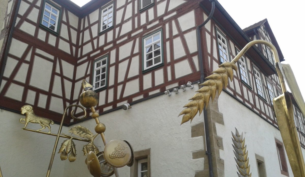 Fachwerkhaus mit goldener Skulptur im Vordergrund, die landwirtschaftliche Symbole wie Getreide und Tiere darstellt., © Natur.Nah. Schönbuch & Heckengäu Fachwerkhaus mit goldener Skulptur im Vordergrund, die landwirtschaftliche Symbole wie Getreide und Tiere darstellt., © Natur.Nah. Schönbuch & Heckengäu