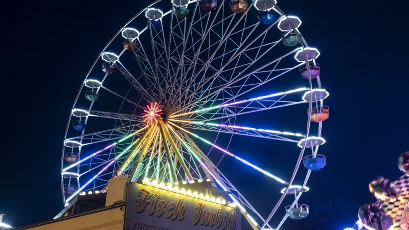 Ein beleuchtetes Riesenrad bei Nacht auf einem Jahrmarkt, mit einem Fisch-Imbiss im Vordergrund., © Torsten Wenzler