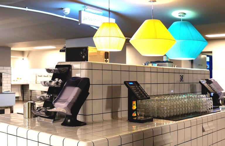 Moderne Frühstückstheke mit bunten Lampen, gestapelten Gläsern und einem Saftspender. Kacheldesign und dezente Beleuchtung., © A&O Hostels Marketing GmbH