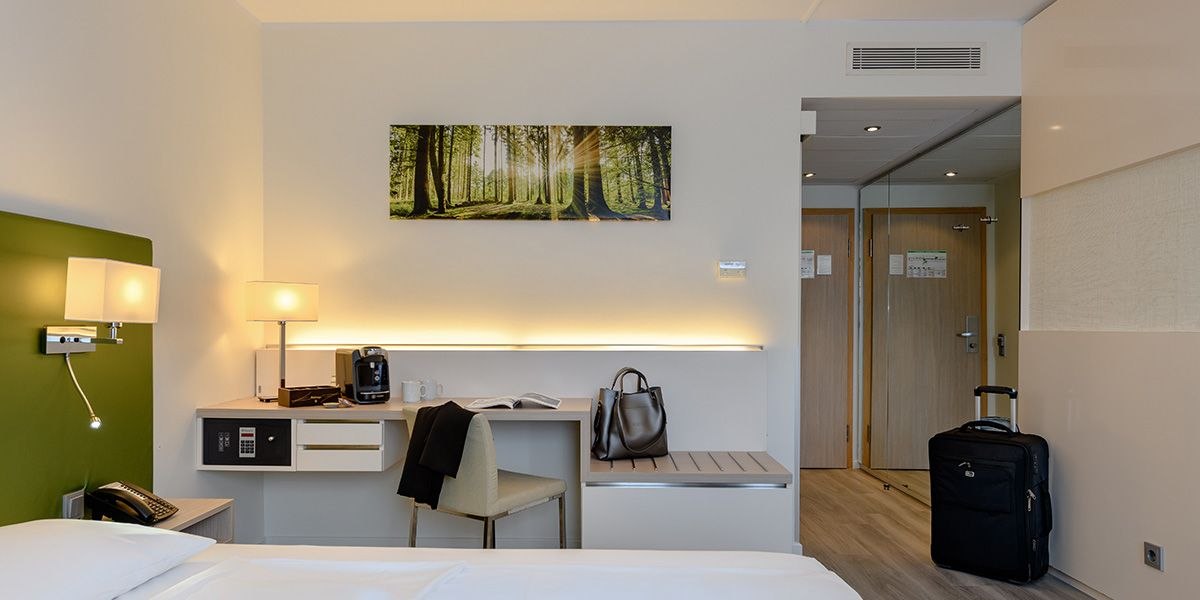 Modernes Hotelzimmer mit Schreibtisch, Koffer, Bild eines Waldes an der Wand und heller Beleuchtung., © Mercure Bristol Stuttgart Sindelfingen