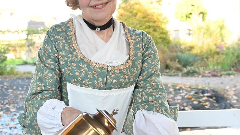 Eine Frau in historischer Kleidung gießt Tee aus einer Kanne in eine Tasse. Sie trägt eine Brille und lächelt. Im Hintergrund ist ein Garten zu sehen., © Copyright: Tourismus & Events Ludwigsburg, Fotograf: Reiner Pfisterer Eine Frau in historischer Kleidung gießt Tee aus einer Kanne in eine Tasse. Sie trägt eine Brille und lächelt. Im Hintergrund ist ein Garten zu sehen., © Copyright: Tourismus & Events Ludwigsburg, Fotograf: Reiner Pfisterer