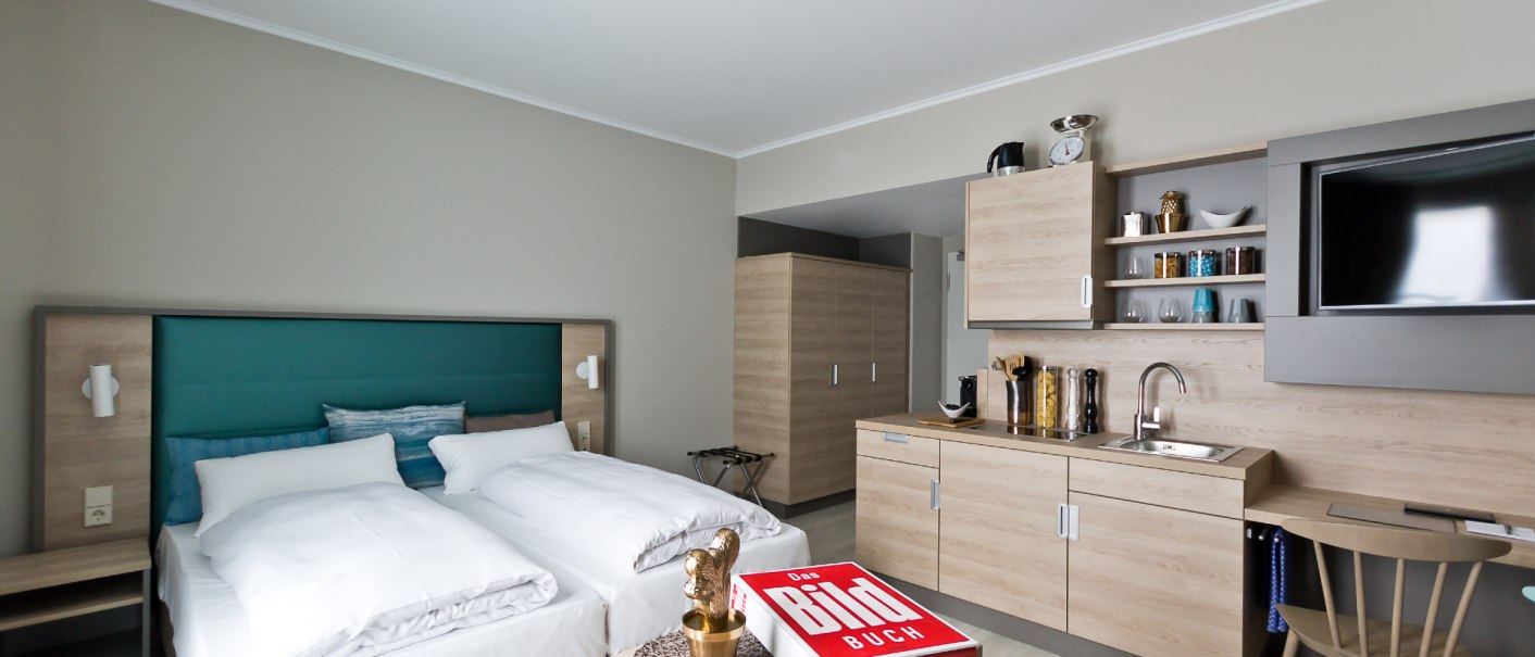 Modernes Apartment mit Doppelbett, Küchenzeile, Tisch und Stuhl. Ein Buch liegt auf dem Tisch, daneben eine goldene Figur., © Arthotel Ana / elaya