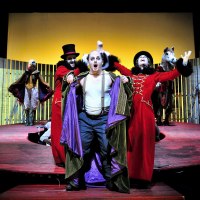 Theaterszene mit Schauspielern in bunten Kostümen auf einer Bühne. Ein Mann in rotem Mantel und Zylinder steht im Vordergrund, andere Figuren im Hintergrund., © Patrick Pfeiffer Theaterszene mit Schauspielern in bunten Kostümen auf einer Bühne. Ein Mann in rotem Mantel und Zylinder steht im Vordergrund, andere Figuren im Hintergrund., © Patrick Pfeiffer