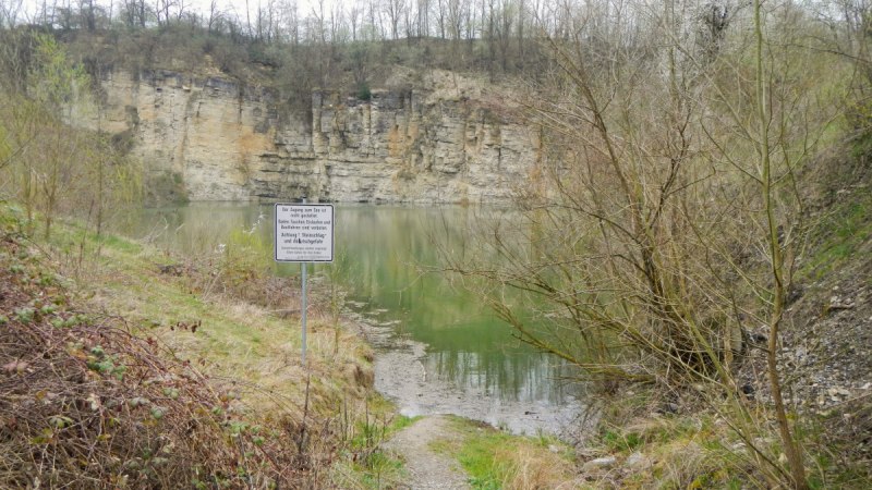 Ein kleiner See mit steilen Felswänden, umgeben von Bäumen. Ein Warnschild weist auf Gefahren hin. Der Himmel ist bewölkt., © Hans Sukowski
