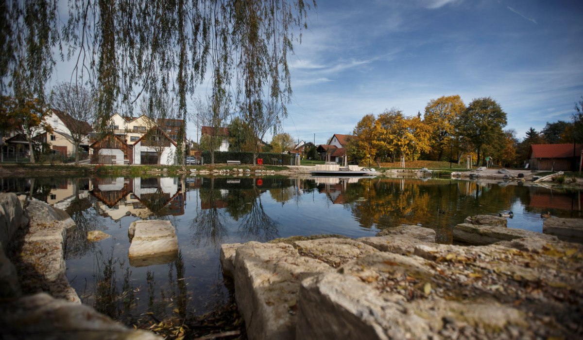 Ein Teich mit Steinen im Vordergrund, umgeben von herbstlichen Bäumen und Häusern. Die ruhige Wasseroberfläche spiegelt die Umgebung wider., © Stoppel Ein Teich mit Steinen im Vordergrund, umgeben von herbstlichen Bäumen und Häusern. Die ruhige Wasseroberfläche spiegelt die Umgebung wider., © Stoppel