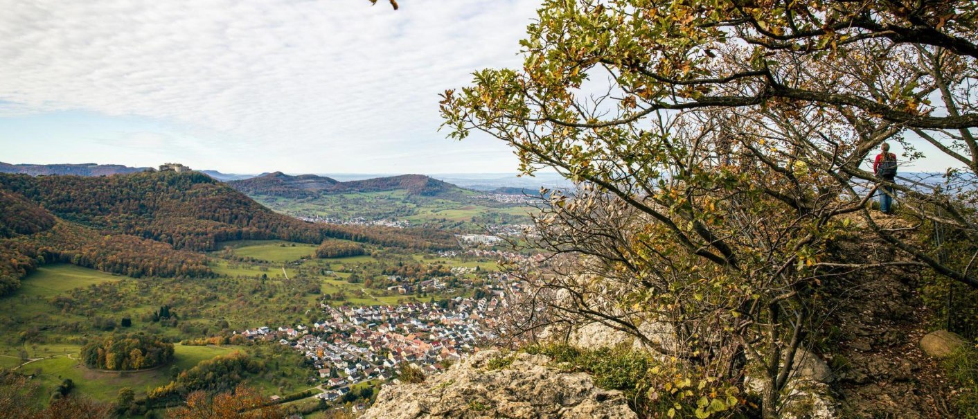 Panoramablick vom Beurener Fels über ein grünes Tal mit einem Dorf und bewaldeten Hügeln im Hintergrund. Ein Wanderer steht am Rand des Felsens., © Stuttgart-Marketing GmbH, Sarah Schmid Panoramablick vom Beurener Fels über ein grünes Tal mit einem Dorf und bewaldeten Hügeln im Hintergrund. Ein Wanderer steht am Rand des Felsens., © Stuttgart-Marketing GmbH, Sarah Schmid