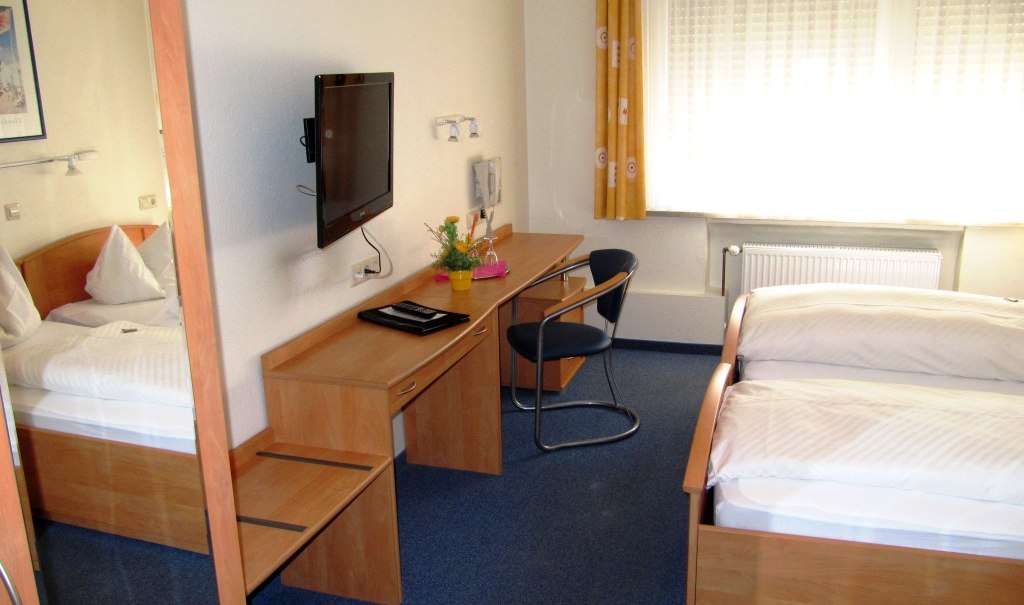 Ein schlichtes Gästezimmer mit zwei Einzelbetten, einem Schreibtisch mit Stuhl, einem Fernseher an der Wand und einem Fenster mit gelben Vorhängen., © R.Storr