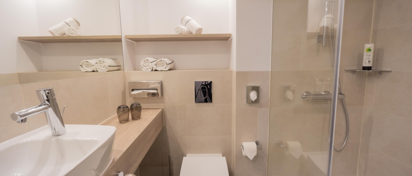 Modernes Badezimmer mit Waschbecken, Toilette, Dusche und Handtüchern auf Regalen. Helle Fliesen und minimalistische Einrichtung., © HARBR. hotel Ludwigsburg / DQuadrat Living GmbH Modernes Badezimmer mit Waschbecken, Toilette, Dusche und Handtüchern auf Regalen. Helle Fliesen und minimalistische Einrichtung., © HARBR. hotel Ludwigsburg / DQuadrat Living GmbH