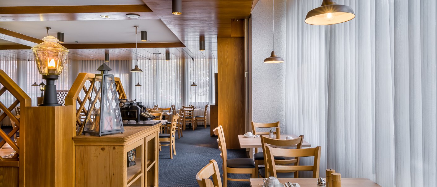 Ein Frühstücksbereich mit Holzmöbeln, gedeckten Tischen und warmem Licht. Große Fenster mit Vorhängen lassen Tageslicht herein., © Novum Management GmbH