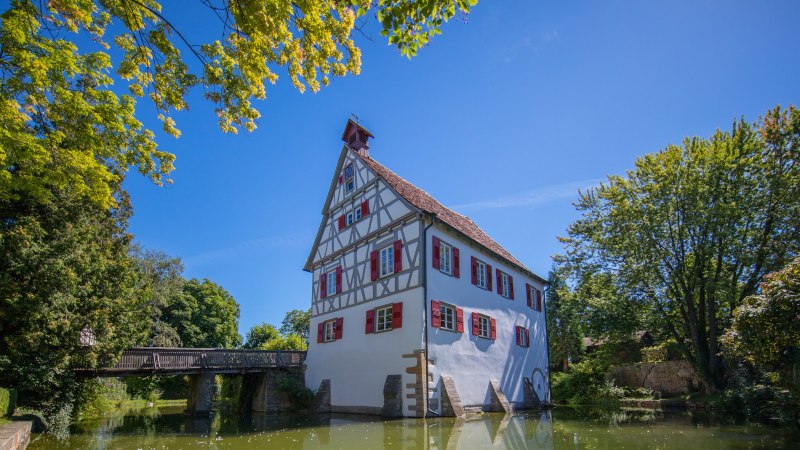 Fachwerkhaus mit roten Fensterläden an einem Teich, umgeben von Bäumen und einer Brücke im Hintergrund, unter blauem Himmel., © SMG, Achim Mende Fachwerkhaus mit roten Fensterläden an einem Teich, umgeben von Bäumen und einer Brücke im Hintergrund, unter blauem Himmel., © SMG, Achim Mende