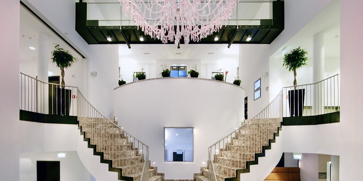 Elegante Lobby mit symmetrischer Treppe, gro&szlig;em Kronleuchter und Pflanzen. Moderne, helle Gestaltung mit Glas und Metallakzenten., &copy; Arcotel Hotels