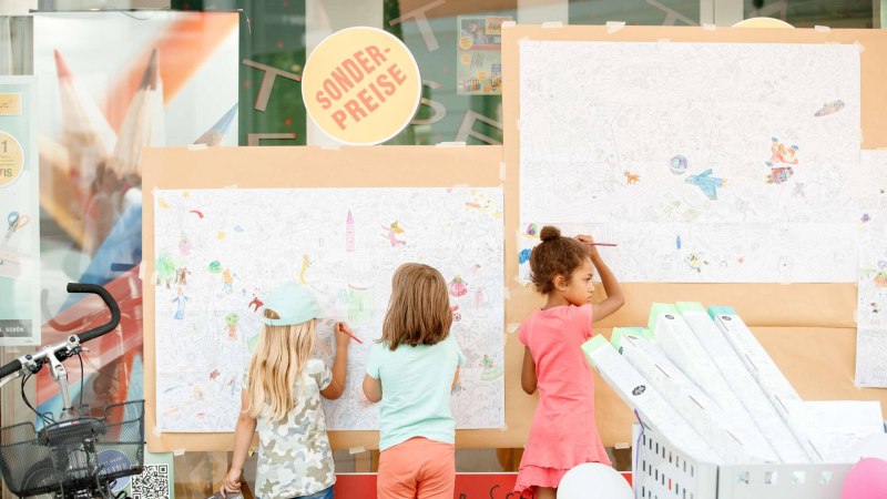 Drei Kinder malen an gro&szlig;en Ausmalpostern vor einem Schaufenster. Ein Sonderpreis-Schild ist sichtbar. Ein Fahrrad steht daneben.