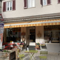 Ein kleiner Laden mit bunten Tischen und Stühlen vor der Tür. Eine Markise spendet Schatten. Im Schaufenster sind Körbe und Dekorationen zu sehen., © L. Besmer