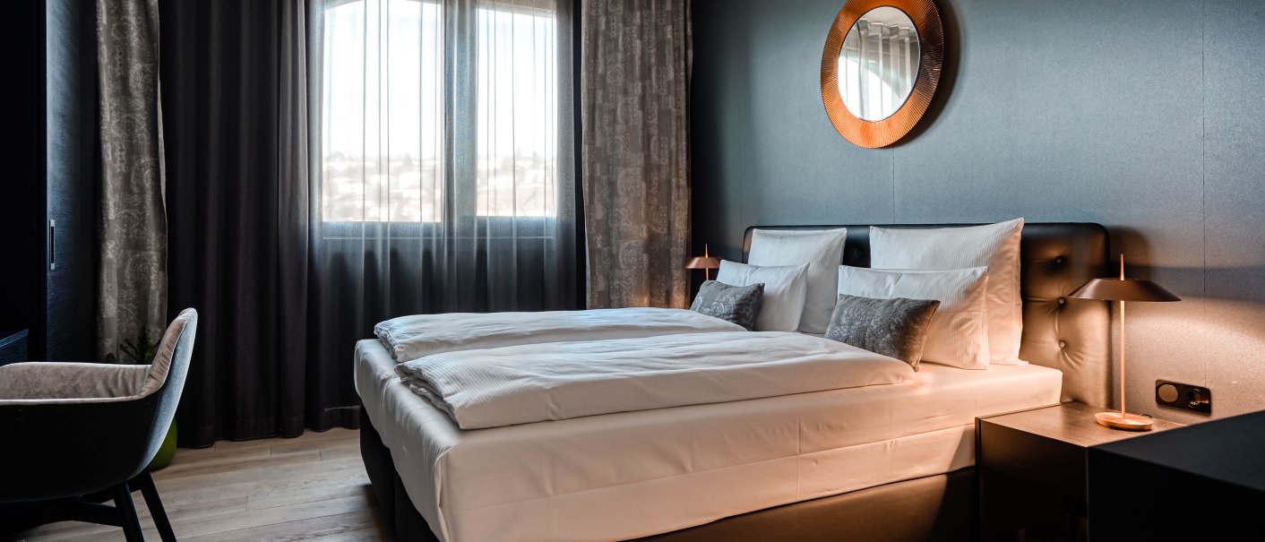 Modernes Schlafzimmer mit Doppelbett, dunklen Vorhängen und einem runden Spiegel an der Wand. Elegante Beleuchtung und Holzboden., © CLOUD N°7 APARTMENTS GmbH