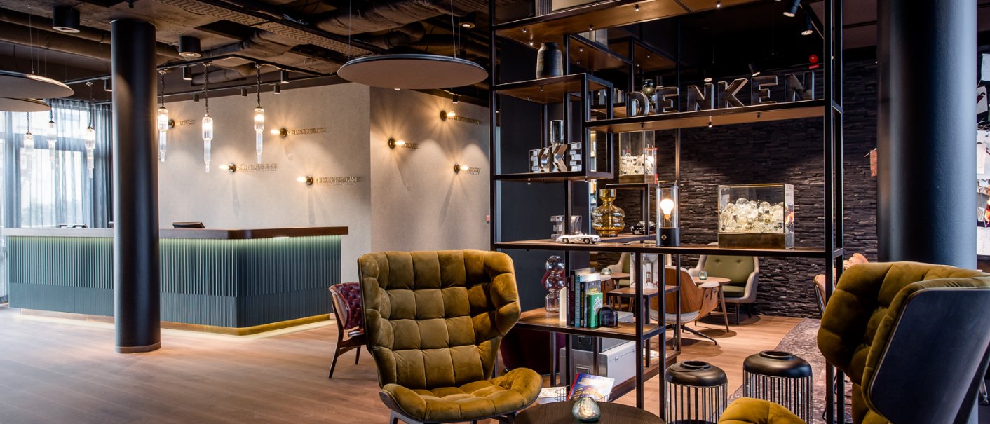 Moderne Hotellobby mit eleganten Sesseln, einem Empfangstresen und stilvoller Beleuchtung. Offenes Regal trennt Sitzbereich ab., © TOMAS