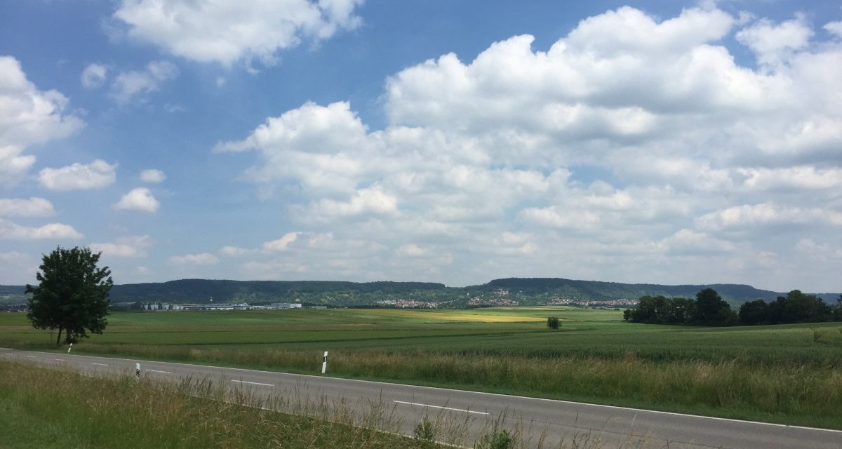 Eine Landstra&szlig;e f&uuml;hrt durch gr&uuml;ne Felder, im Hintergrund sind H&uuml;gel und ein bew&ouml;lkter Himmel zu sehen., &copy; www.pro-cycl.de