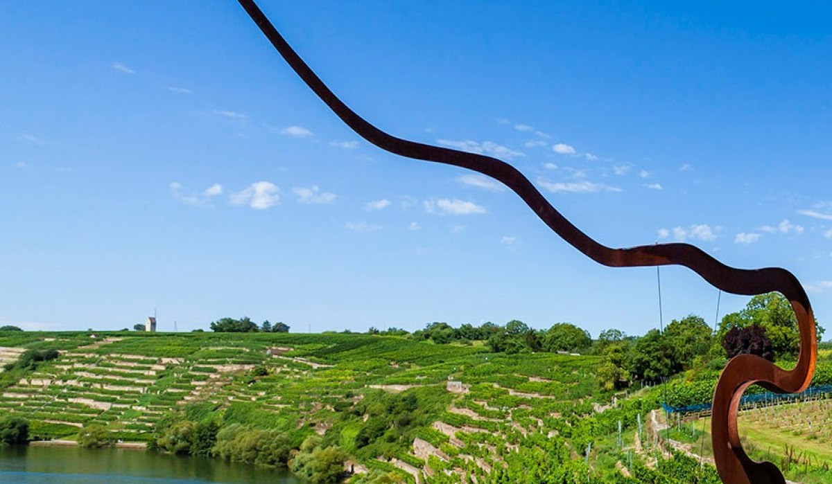 Weinberge mit einer geschwungenen Skulptur im Vordergrund, ein See und blauer Himmel im Hintergrund., © Land der 1000 Hügel - Kraichgau-Stromberg Weinberge mit einer geschwungenen Skulptur im Vordergrund, ein See und blauer Himmel im Hintergrund., © Land der 1000 Hügel - Kraichgau-Stromberg