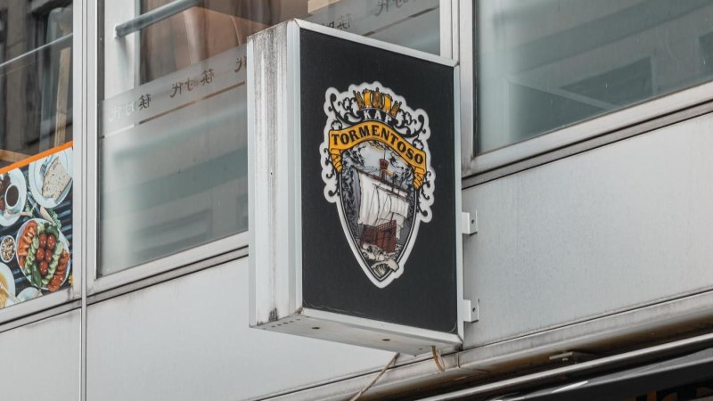 Ein Schild mit dem Logo von Kap Tormentoso hängt an einem modernen Gebäude. Es zeigt ein Segelschiff und dekorative Elemente., © Stuttgart Marketing GmbH, Sarah Schmid Ein Schild mit dem Logo von Kap Tormentoso hängt an einem modernen Gebäude. Es zeigt ein Segelschiff und dekorative Elemente., © Stuttgart Marketing GmbH, Sarah Schmid