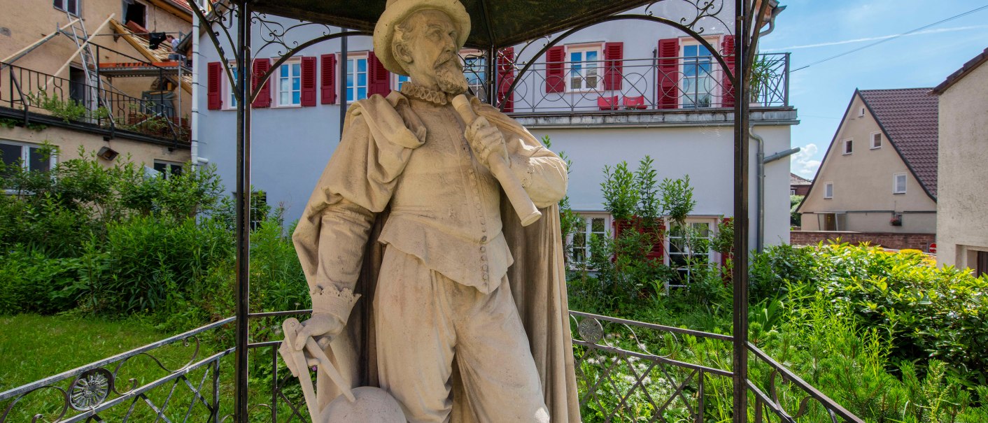 Steinstatue eines Mannes mit Hut und Umhang, der ein Instrument hält. Sie steht unter einem Pavillon in einem grünen Garten, umgeben von Häusern., © Stuttgart-Marketing GmbH, Achim Mende