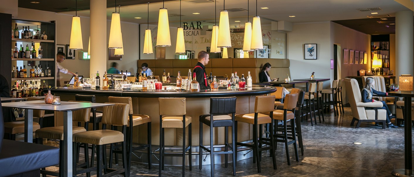 Moderne Bar mit runder Theke, Hängelampen und Sitzbereichen im Arcotel Camino. Menschen sitzen und genießen die Atmosphäre., © Harald Eisenberger
