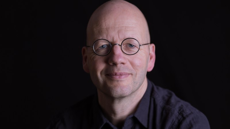 Ein Mann mit runder Brille und Glatze lächelt leicht in die Kamera. Der Hintergrund ist schwarz, was den Fokus auf sein Gesicht lenkt., © Frank Suffert