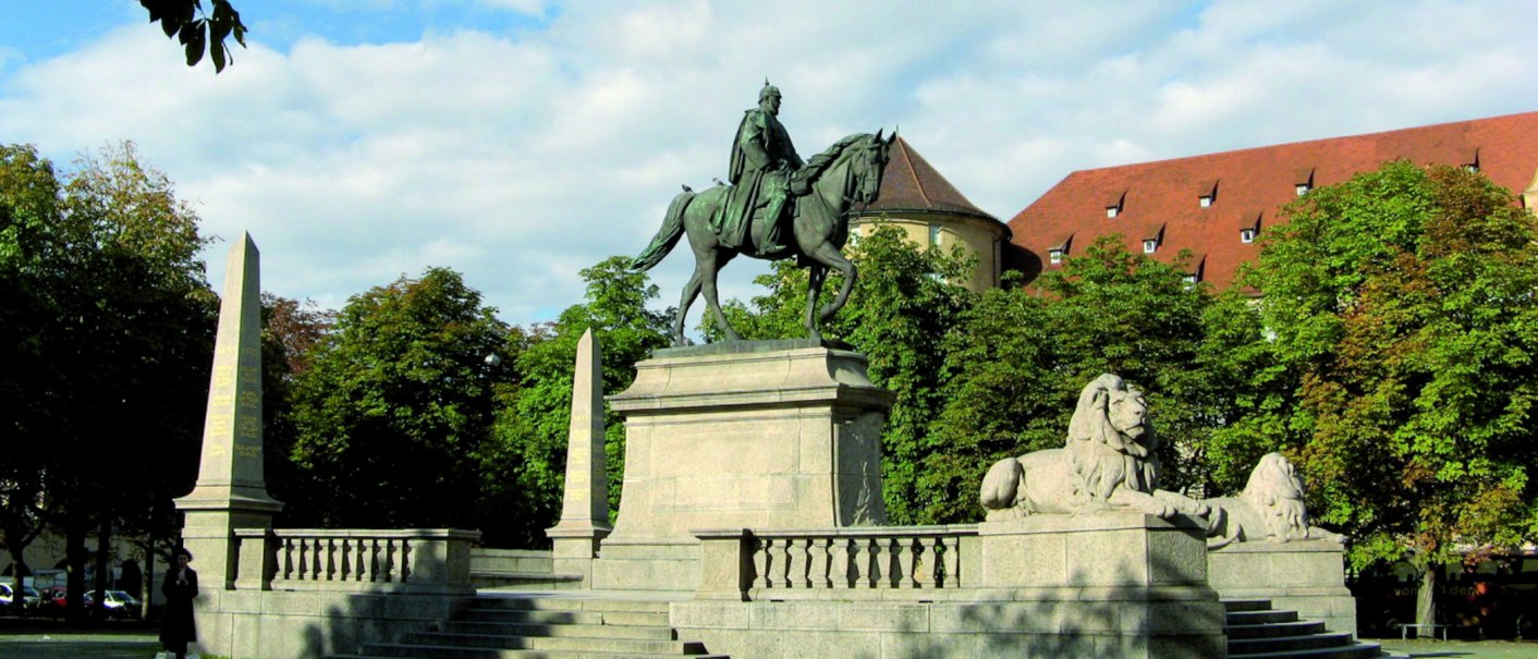 Reiterstatue auf dem Karlsplatz in Stuttgart, flankiert von Löwenstatuen und Obelisken, umgeben von Bäumen und einem historischen Gebäude im Hintergrund., © Stuttgart-Marketing GmbH Reiterstatue auf dem Karlsplatz in Stuttgart, flankiert von Löwenstatuen und Obelisken, umgeben von Bäumen und einem historischen Gebäude im Hintergrund., © Stuttgart-Marketing GmbH