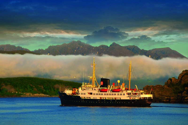 Ein Hurtigruten-Schiff fährt auf ruhigem Wasser, umgeben von Bergen und Wolken. Die Szene wirkt friedlich und malerisch., © BLICKFANG Ein Hurtigruten-Schiff fährt auf ruhigem Wasser, umgeben von Bergen und Wolken. Die Szene wirkt friedlich und malerisch., © BLICKFANG