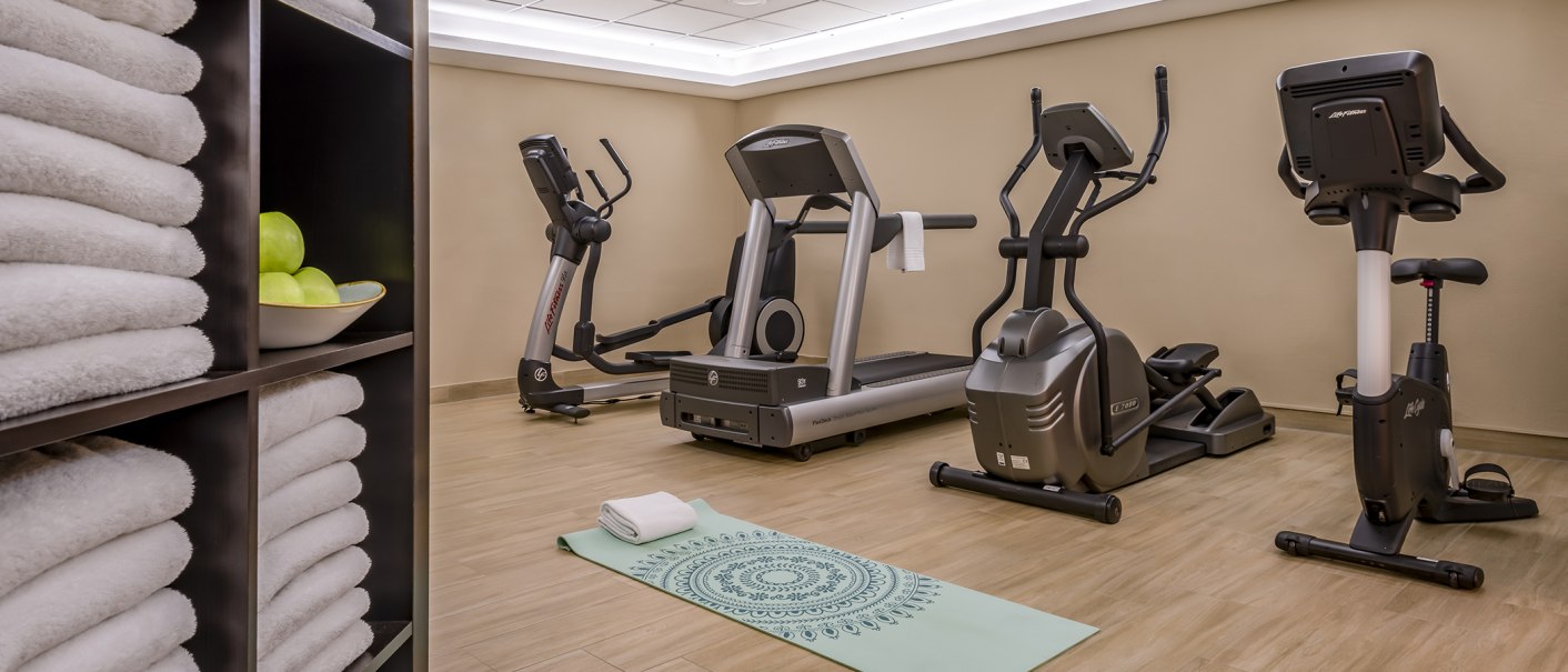 Fitnessraum mit Laufband, Crosstrainer, Heimtrainer, Yogamatte und Handtüchern. Ein Regal mit Handtüchern und Äpfeln steht im Vordergrund., © Stuttgart Marriott Hotel Sindelfingen
