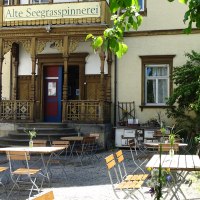 Die Alte Seegrasspinnerei mit verzierten Holzdetails und einem Außenbereich mit Tischen und Stühlen unter Bäumen., © Alte Seegrasspinnerei