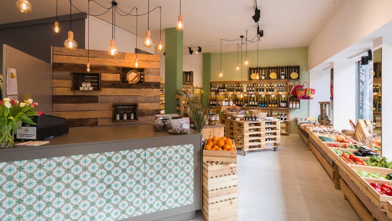 Moderner Feinkostladen mit Holzregalen, frischem Obst und Gem&uuml;se. H&auml;ngelampen und eine Theke mit dekorativen Fliesen verleihen eine gem&uuml;tliche Atmosph&auml;re., &copy; Benjamin Stollenberg