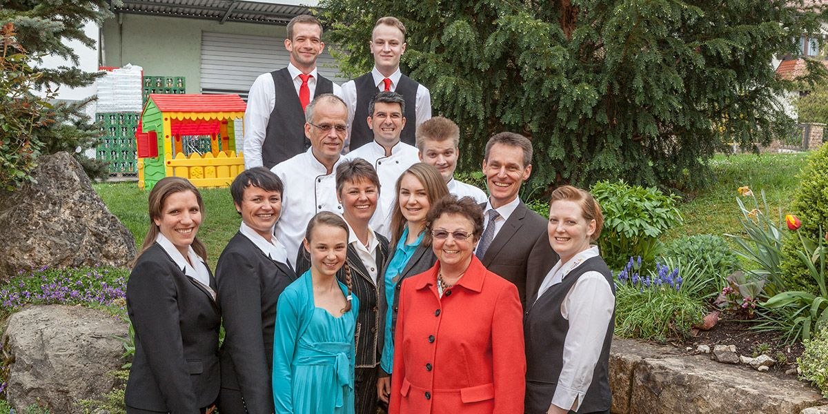 Eine Gruppe von Menschen in formeller Kleidung posiert lächelnd im Freien vor einem Garten mit Bäumen und Blumen., © Hotel Restaurant Lamm