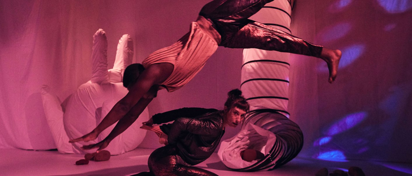 Zwei Akrobaten in einem pink beleuchteten Raum. Einer springt elegant über den anderen, der in einer gebückten Position verharrt., © JES, Alex Wunsch