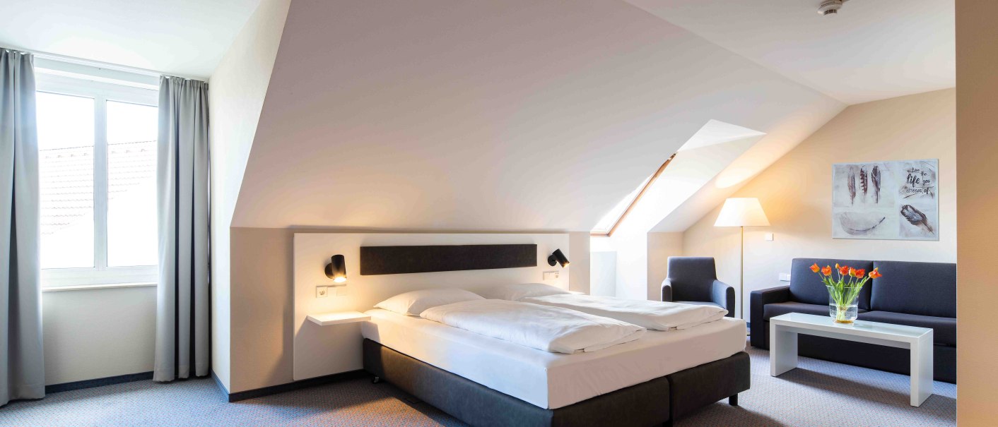 Helles Hotelzimmer mit Doppelbett, Sofa, Tisch und Blumen. Schräge Decke, Fenster mit Vorhängen, moderne Einrichtung., © Erikson Hotel Helles Hotelzimmer mit Doppelbett, Sofa, Tisch und Blumen. Schräge Decke, Fenster mit Vorhängen, moderne Einrichtung., © Erikson Hotel