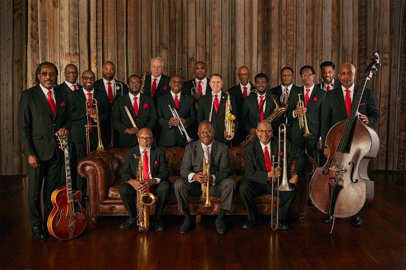 Eine Jazzband in Anzügen mit roten Krawatten posiert mit Instrumenten vor einer Holzwand., © Backnang