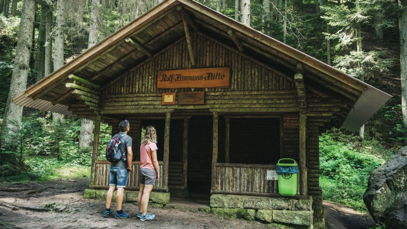 Zwei Personen betrachten die Rolf Hamann Hütte im Wald. Die Hütte ist aus Holz und von Bäumen umgeben., © LOCHER Fotodesign&Manufaktur