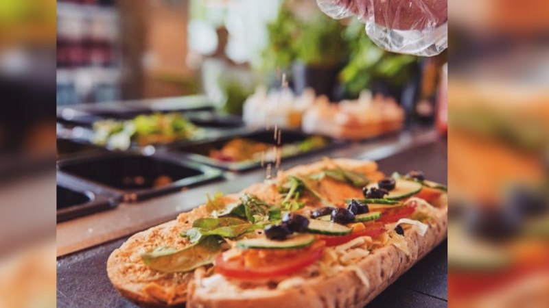 Ein Sandwich wird mit frischem Gemüse und Oliven belegt. Eine Hand streut Gewürze darüber. Im Hintergrund sind Zutaten in Behältern zu sehen., © FreshSub Ein Sandwich wird mit frischem Gemüse und Oliven belegt. Eine Hand streut Gewürze darüber. Im Hintergrund sind Zutaten in Behältern zu sehen., © FreshSub