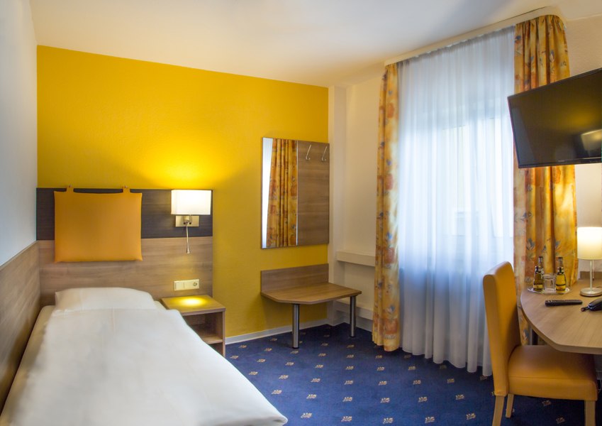 Ein gemütliches Einzelzimmer mit gelber Akzentwand, Einzelbett, Schreibtisch, Stuhl und Fernseher. Helle Vorhänge und Teppichboden ergänzen das Ambiente., © Römerhof Hotelbetriebs GmbH