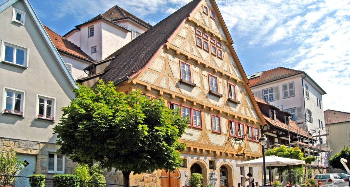 Fachwerkhaus in einer Altstadt mit Caf&eacute; und Sonnenschirmen. Menschen sitzen drau&szlig;en, ein Baum spendet Schatten. Der Himmel ist blau mit wenigen Wolken., &copy; Graphic Design Albig, GDA Waldenbuch