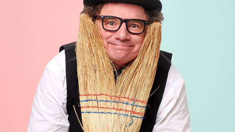 Ein Mann mit Zylinder und Brille posiert humorvoll mit einem Besen als Bart vor einem zweifarbigen Hintergrund in Rosa und Hellblau.