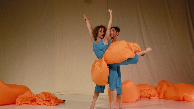 Zwei T&auml;nzer in blauen Kost&uuml;men posieren mit orangefarbenen Kissen auf einer B&uuml;hne. Der Hintergrund ist beige, eine Discokugel h&auml;ngt oben., &copy; FILharmonie Filderstadt