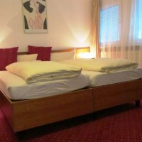 Hotelzimmer mit Doppelbett, rotem Teppich, Nachttischlampe und Bild an der Wand. Fenster mit Vorhängen, Tisch mit Vase., © TOMAS