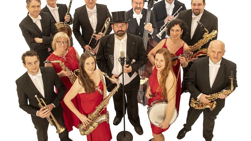 Eine Musikgruppe in eleganter Kleidung posiert mit verschiedenen Instrumenten. Die M&auml;nner tragen Anz&uuml;ge, die Frauen rote Kleider. Sie stehen vor einem wei&szlig;en Hintergrund., &copy; Touristik Bad Wildbad GmbH